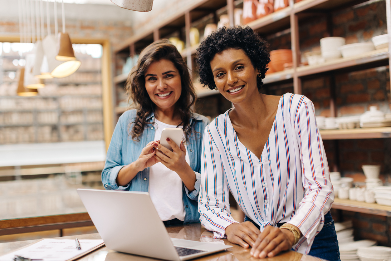 Happy ceramists smiling at the camera while working with wireless technology. Female entrepreneurs managing online orders in their store. Two businesswomen running a creative small business together.