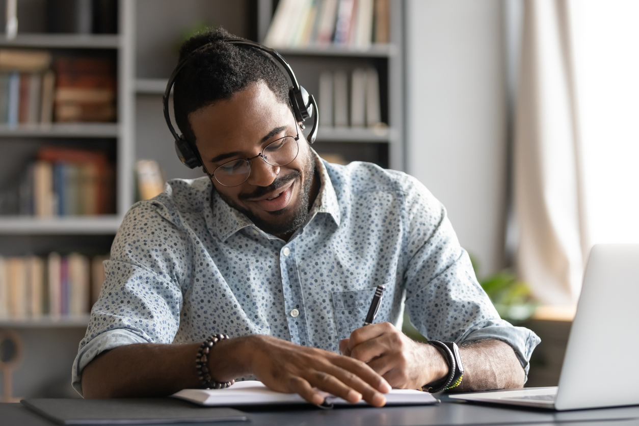 Smiling african male student professional wear headset elearning study with online tutor teacher listening audio podcast e learning language concept make notes sit at home office desk with laptop