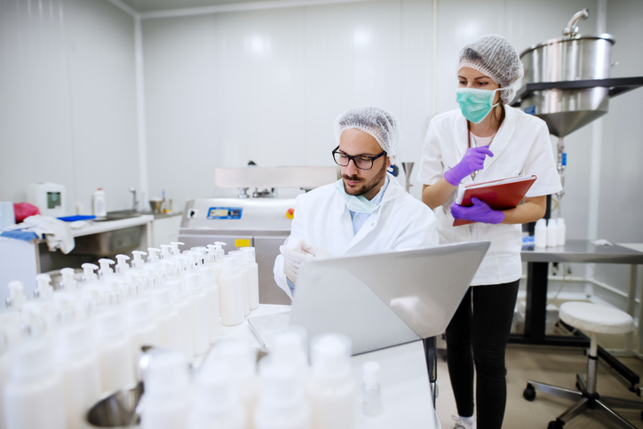 scientist doing research in cosmetic factory. next to him assistant with folder in hands. on the desk bottles and laptop.