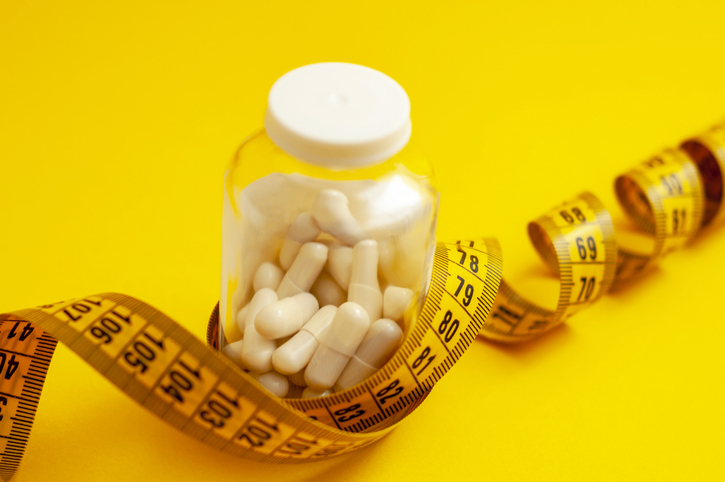 tablets in capsules in a glass jar, tied with measuring tape on a yellow background, close up, horizontal shot. the concept of weight loss, fat burner, vitamins, sports nutrition.