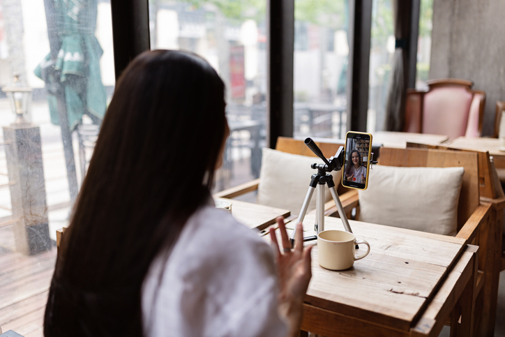 happy young caucasian millennial or gen z woman with long brunette hair streaming with smart phone on tripod, shooting social media blog in modern cafe. influencer using social networks indoor